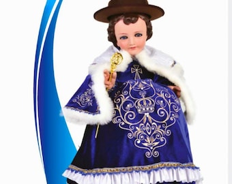 Puede incluir: Una figura de un niño que lleva un vestido azul y blanco con bordados dorados y una corona. El niño lleva un sombrero marrón y sostiene un cetro dorado. La figura está titulada "NIÑO DE ATOCHA".