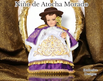 Puede incluir: Una figura de un niño vestido con una túnica blanca con bordados dorados y una capa morada. El niño lleva un sombrero marrón y sostiene un cetro dorado. La figura está sentada en un trono dorado.