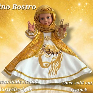 May include: A doll dressed in a white and gold embroidered gown, holding a gold cross. The doll has a gold head covering and a gold scarf. The text "Divino Rostro" is at the top of the image.
