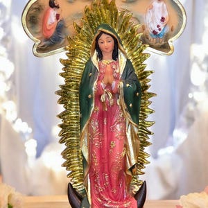 May include: A religious figurine of a woman in a pink and green robe, with a gold halo and cross-shaped backdrop. The figurine is standing on a base with a child and surrounded by roses. The text "NlartesaniaMexicanaExclusiveDesigns" is at the bottom.