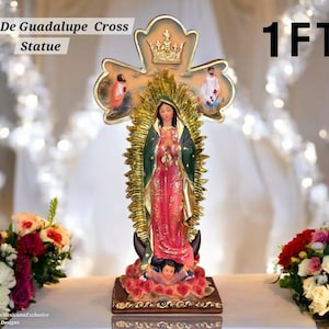 May include: A statue of the Virgin of Guadalupe, approximately 1 foot tall, stands before a decorative cross. The statue features a figure in a pink robe and green cloak, with a gold halo. The cross has gold accents and small figures.