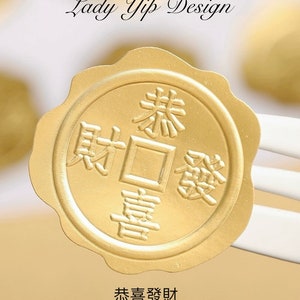 Könnte beinhalten: Ein runder goldener Aufkleber mit chinesischen Schriftzeichen, die sich mit "Ich wünsche dir Wohlstand" übersetzen lassen. Der Aufkleber hat ein erhabenes, geprägtes Design und einen gewellten Rand.