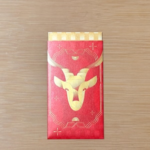 May include: A rectangular red envelope featuring a gold foil illustration of an antelope head centered within a geometric frame. The top flap displays a gold and white checkered pattern, while small gold cross symbols are scattered across the red surface.