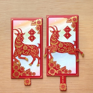 May include: Two rectangular red envelopes featuring a gold and red paper-cut style goat illustration. The designs include floral patterns, a diamond-shaped emblem with Chinese characters, and a small tab at the bottom with additional text.