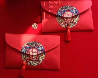 May include: Two red envelopes with gold tassels and intricate embroidered designs. The envelopes feature a circular design with the Chinese character for "double happiness" in the center.