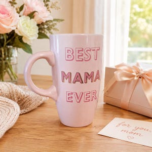 Może przedstawiać: Jasnoróżowy ceramiczny kubek z napisem "BEST MAMA EVER" w kolorze różowym. Kubek ma zakrzywiony uchwyt i stoi na drewnianej powierzchni. Obecne jest również pudełko prezentowe z wstążką i kartka z napisem "for you, mom".