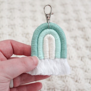 May include: A light blue and white rainbow keychain with a silver key ring. The rainbow is made of yarn and has a white fringe.