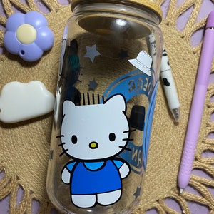 May include: A clear glass water bottle with a wooden lid and a Hello Kitty design. The bottle has a blue and white design with stars and the text "Sayan".