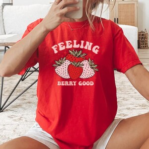 Può includere: Una maglietta rossa con le parole "Feeling Berry Good" e una grafica di fragole. La maglietta è a maniche corte e ha un girocollo. La persona indossa pantaloncini bianchi e calzini bianchi con strisce nere.