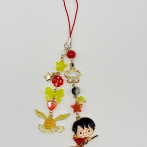 May include: A decorative phone charm featuring a red cord, gold-tone metal charms of a winged ball with the text 'I open at the close' and a cartoon character on a broomstick, accented with yellow stars, bows, red beads, and a pearl-like flower.