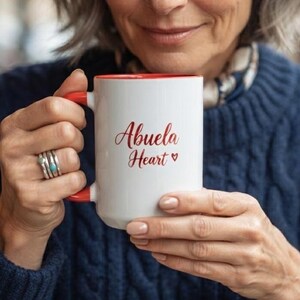 May include: A white ceramic mug with a red interior and a red handle. The exterior features the red text Abuela Heart with a small heart symbol. The mug is held by two hands, one of which wears a silver ring with a small blue stone on the finger.
