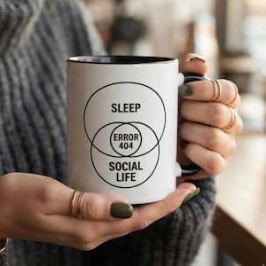Può includere: Una tazza in ceramica bianca con interno e manico neri, caratterizzata da un diagramma di Venn nero. Il diagramma mostra il testo SLEEP nel cerchio superiore, SOCIAL LIFE nel cerchio inferiore e ERROR 404 nella sezione centrale sovrapposta.