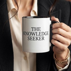 May include: A white ceramic mug with a black interior and handle, featuring the black text THE KNOWLEDGE SEEKER printed in a serif font on the front. The mug has a cylindrical shape and a smooth finish, held by a handle on the right side.