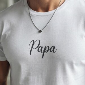 May include: A white short-sleeved t-shirt featuring the word Papa printed in a dark gray cursive script across the chest. A thin silver-toned metal chain necklace with a small geometric pendant rests against the neckline of the shirt.