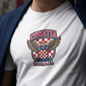 Puede incluir: Una camiseta blanca de manga corta con un dise&ntilde;o gr&aacute;fico que incluye el texto CROATIA en la parte superior y FOOTBALL en la inferior. El emblema central muestra un &aacute;guila dorada con las alas extendidas sobre un escudo ajedrezado.