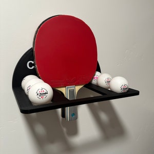 May include: A black wall-mounted rack with a red and black ping pong paddle and five white ping pong balls. The balls have a red star and the text "Nittaku Premium 40+" printed on them.