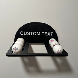 May include: Black wall-mounted ping pong ball holder with the text "CUSTOM TEXT" on the front. The holder has six slots for ping pong balls, and there are five white ping pong balls in the slots. The balls have a pink and white logo.