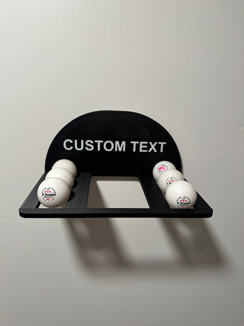 May include: Black wall-mounted ping pong ball holder with the text "CUSTOM TEXT" on the front. The holder has six slots for ping pong balls, and there are five white ping pong balls in the slots. The balls have a pink and white logo.