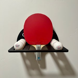 May include: A black wall-mounted rack with a red and black ping pong paddle and six white ping pong balls. The balls have the text "Nittaku 40+" printed on them.