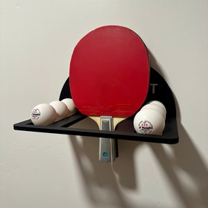 May include: A black wall-mounted rack with a red and black ping pong paddle and six white ping pong balls. The rack has a small black sign that says "PING PONG".