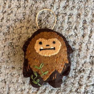 Può includere: Ornamento fatto a mano in feltro a forma di Bigfoot. L'ornamento presenta feltro marrone e beige con dettagli ricamati, tra cui un viso, pelliccia e un rametto di foglie verdi. Un anello di spago è attaccato per appenderlo.