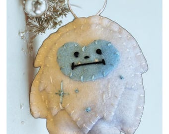 Puede incluir: Adorno de fieltro hecho a mano con forma de yeti amigable. El yeti tiene una cara azul claro, ojos y boca negros cosidos, y está adornado con pequeñas cuentas azules e hilo plateado. Se adjunta un lazo plateado para colgarlo.