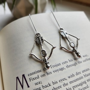 May include: Silver-coloured earrings in the shape of bows and arrows. Each earring features an arrow, a central stone, and a heart-shaped design. The earrings are presented on an open book with printed text.