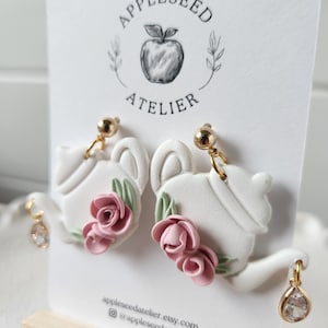 May include: White teapot shaped earrings with pink roses and green leaves. The earrings have gold posts and crystal accents.