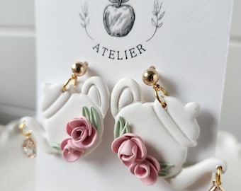 May include: White teapot shaped earrings with pink roses and green leaves. The earrings have gold posts and crystal accents.