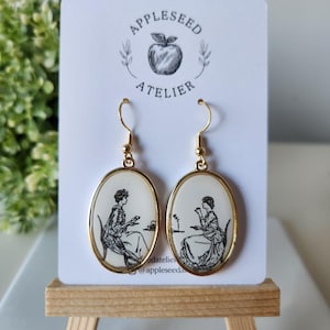 Puede incluir: Pendientes ovalados dorados con una ilustración en blanco y negro de dos mujeres sentadas tomando el té. Los pendientes están en una tarjeta blanca con el texto "Appleseed Atelier".