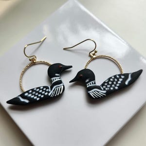 May include: A pair of dangle earrings featuring black and white loon bird charms suspended from thin, gold-toned textured circular hoops with standard hook-style ear wires for secure attachment to the earlobe.