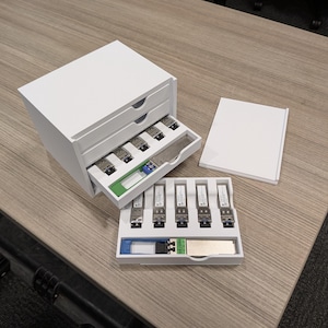 Może przedstawiać: Białe, dwuszeregowe pudełko do przechowywania z otwartymi szufladami, ujawniające kilka srebrnych i czarnych transceiverów sieciowych. Taca z dodatkowymi transceiverami i stos białego papieru również znajdują się na drewnianej powierzchni.