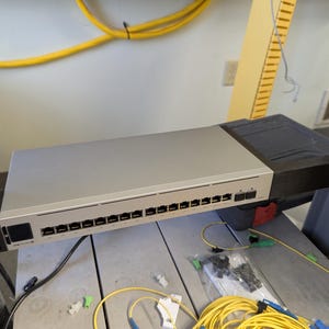 Puede incluir: Un conmutador de red plateado con 16 puertos montado en un rack. El conmutador está conectado a un haz de cables de fibra óptica amarillos.