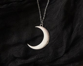 May include: A silver crescent moon pendant necklace on a black fabric background.