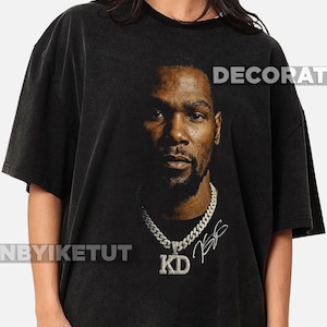 May include: Black t-shirt featuring a portrait of a man with a diamond-studded chain and pendant. The pendant reads "KD". The shirt has the word "DECORAT" printed on the top right and "NBYIKETUT" on the bottom left.
