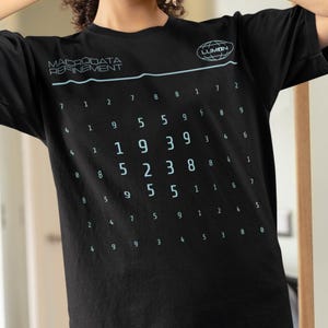 Może przedstawiać: Czarny T-shirt z białym nadrukiem graficznym z tekstem "MACRODATA REFINEMENT" i siatką liczb. Grafika zawiera również małe kółko z tekstem "LUMON" w środku.