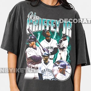 May include: A charcoal gray short-sleeve t-shirt featuring a graphic print of Ken Griffey Jr. in various baseball poses. The design includes the text "Ken Griffey Jr." in a stylized font, surrounded by bright teal lightning bolts on a dark background.