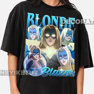 May include: Black t-shirt with a graphic of a blonde woman wearing a blue mask and the text "BLONDE BLAZER". The design features multiple images of the character in a comic book style.