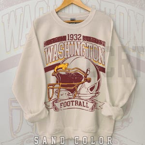 May include: Sand-coloured sweatshirt with a distressed graphic featuring a football helmet and the text "WASHINGTON FOOTBALL 1932." The design includes burgundy and gold accents. The sweatshirt is displayed on a wooden hanger.