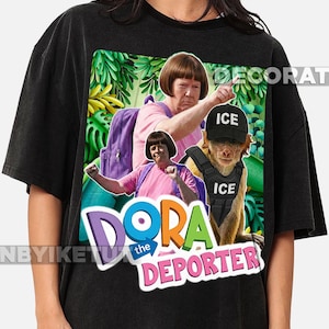 Op de afbeelding: Zwart t-shirt met een parodie-ontwerp. De afbeelding toont de tekst "Dora the Deporter" en een humoristische weergave van Dora the Explorer met een aap die een "ICE" vest en hoed draagt, tegen een jungle achtergrond.