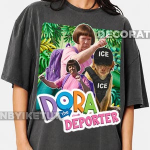 May include: A charcoal grey t-shirt featuring a graphic print of a person with a brown bob haircut wearing a pink shirt and purple backpack, alongside a monkey wearing a black cap and vest labelled ICE, with the text DORA the DEPORTER.