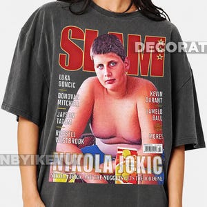 May include: A charcoal grey short-sleeve t-shirt featuring a graphic print of a vintage SLAM magazine cover. The cover displays a young Nikola Jokic with the text "NIKOLA JOKIC" and "NUGGETS GET THE JOB DONE" along with various player names.