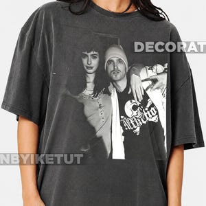 Könnte beinhalten: Ein anthrazitfarbenes Kurzarm-T-Shirt mit einem schwarz-weißen Fotodruck von zwei Personen. Die Person links hat dunkles Haar, die Person rechts trägt eine Mütze und ein T-Shirt mit einem Totenkopf-Grafikdesign auf der Vorderseite.