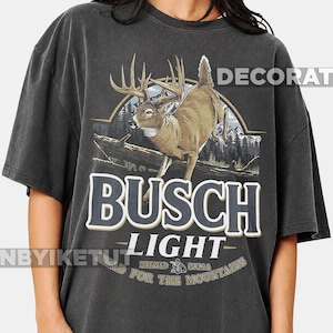 May include: A charcoal gray short-sleeve t-shirt featuring a graphic of a deer jumping over a log in front of mountains. The text reads BUSCH LIGHT, BREWED IN USA, and HEAD FOR THE MOUNTAINS. The design includes a circular border and forest imagery.