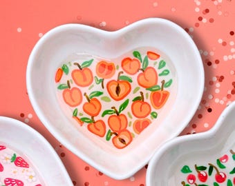 May include: A white heart-shaped dish with a peach design. The dish is filled with a clear liquid and has a peach pattern on the bottom.