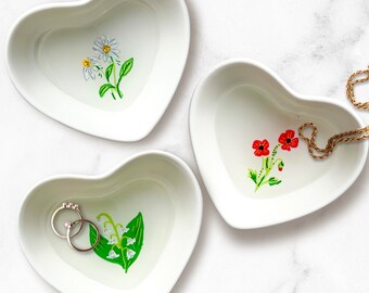 May include: Three white heart-shaped ceramic dishes. Each dish features a hand-painted floral design: daisies, red poppies, and lily of the valley. Two rings rest in one dish, and a gold necklace in another. Ideal for jewelry or small items.