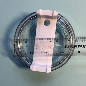 May include: A clear, round lid with a white plastic slider, measuring approximately 4 inches in diameter. A ruler is placed across the lid, showing measurements in inches and centimeters. The lid is on a light blue surface.