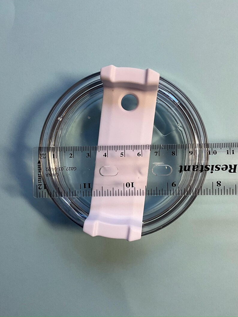 May include: A clear, round lid with a white plastic slider, measuring approximately 4 inches in diameter. A ruler is placed across the lid, showing measurements in inches and centimeters. The lid is on a light blue surface.