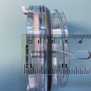 May include: A clear plastic container with a black rubber seal, measured with a ruler. The ruler shows the container is approximately 1.2 inches in diameter. The text "CHINA G027 03-04/22" is visible on the ruler.