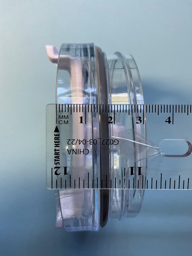 May include: A clear plastic container with a black rubber seal, measured with a ruler. The ruler shows the container is approximately 1.2 inches in diameter. The text "CHINA G027 03-04/22" is visible on the ruler.